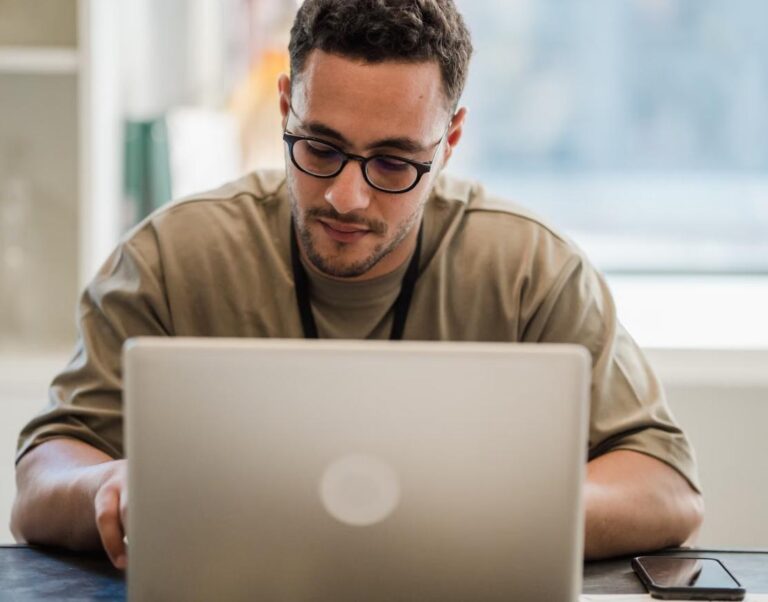 A person working focused on a laptop at a home desk near a window.