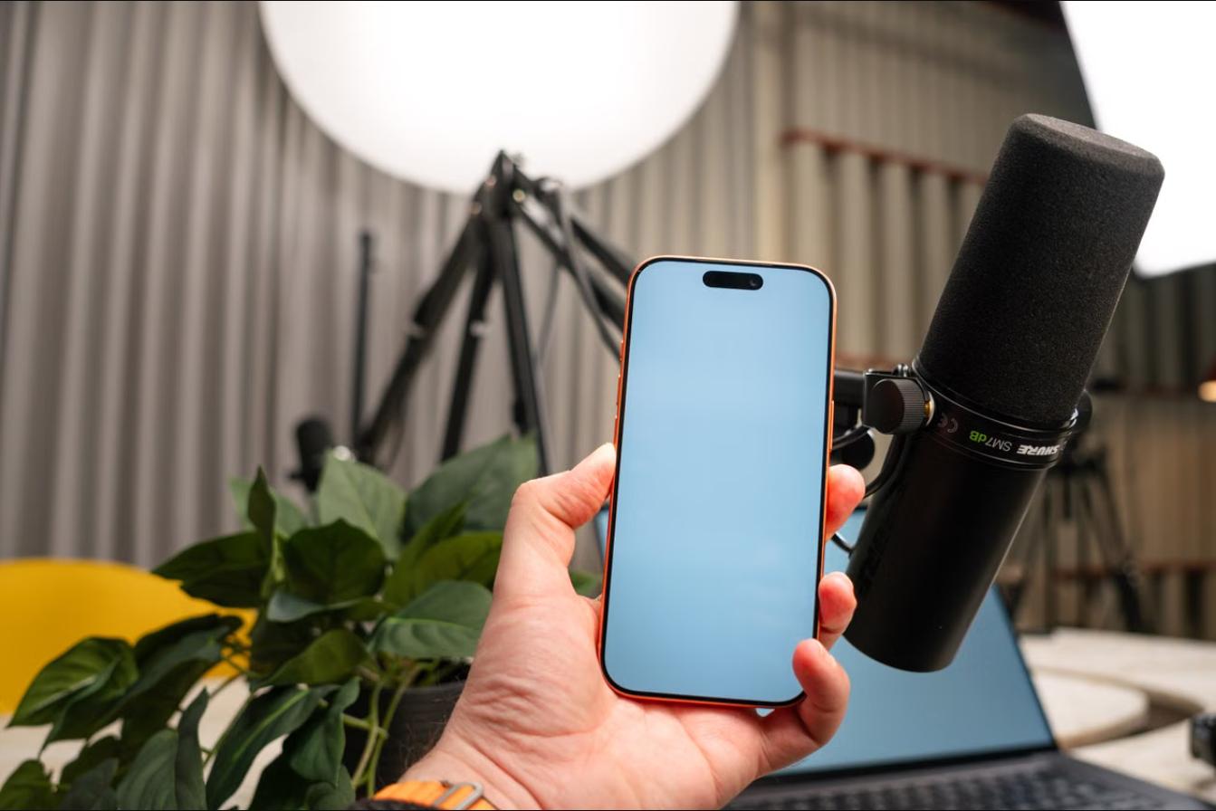 A hand holding a smartphone next to a podcast microphone for audio recording.