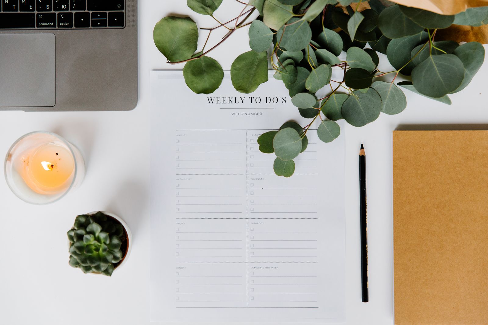 Flat lay of a desk with a laptop, a weekly to-do list planner, a notebook, a candle and eucalyptus leaves