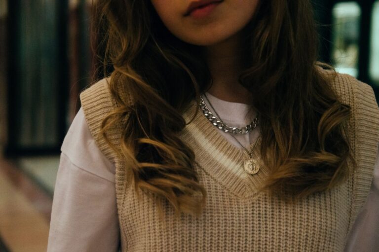 Beige knit sweater vest layered over a white shirt with a silver chain and coin pendant necklace