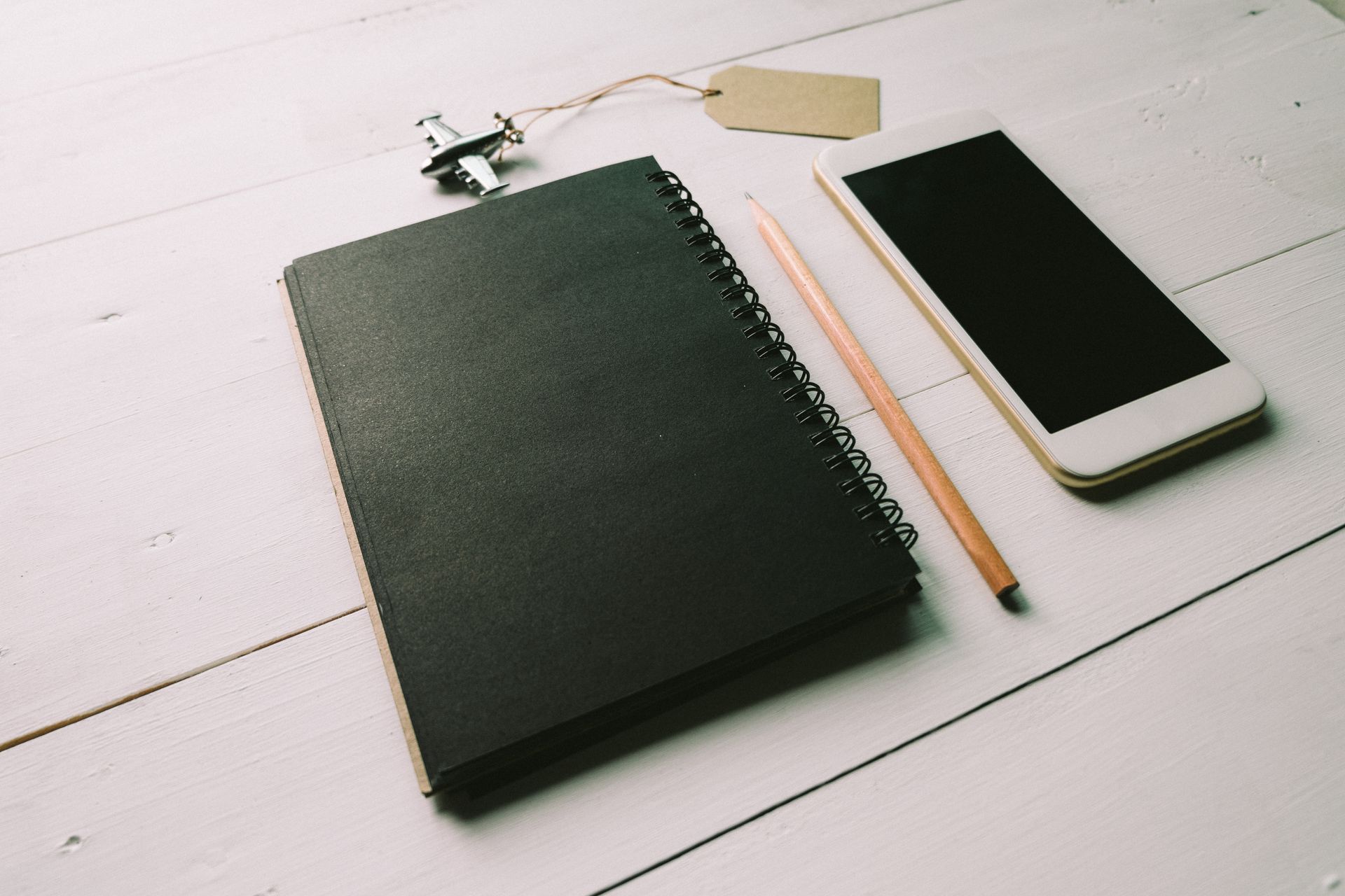 Flat lay of a black spiral notebook, a pencil and a smartphone with a dark screen on a white wooden desk