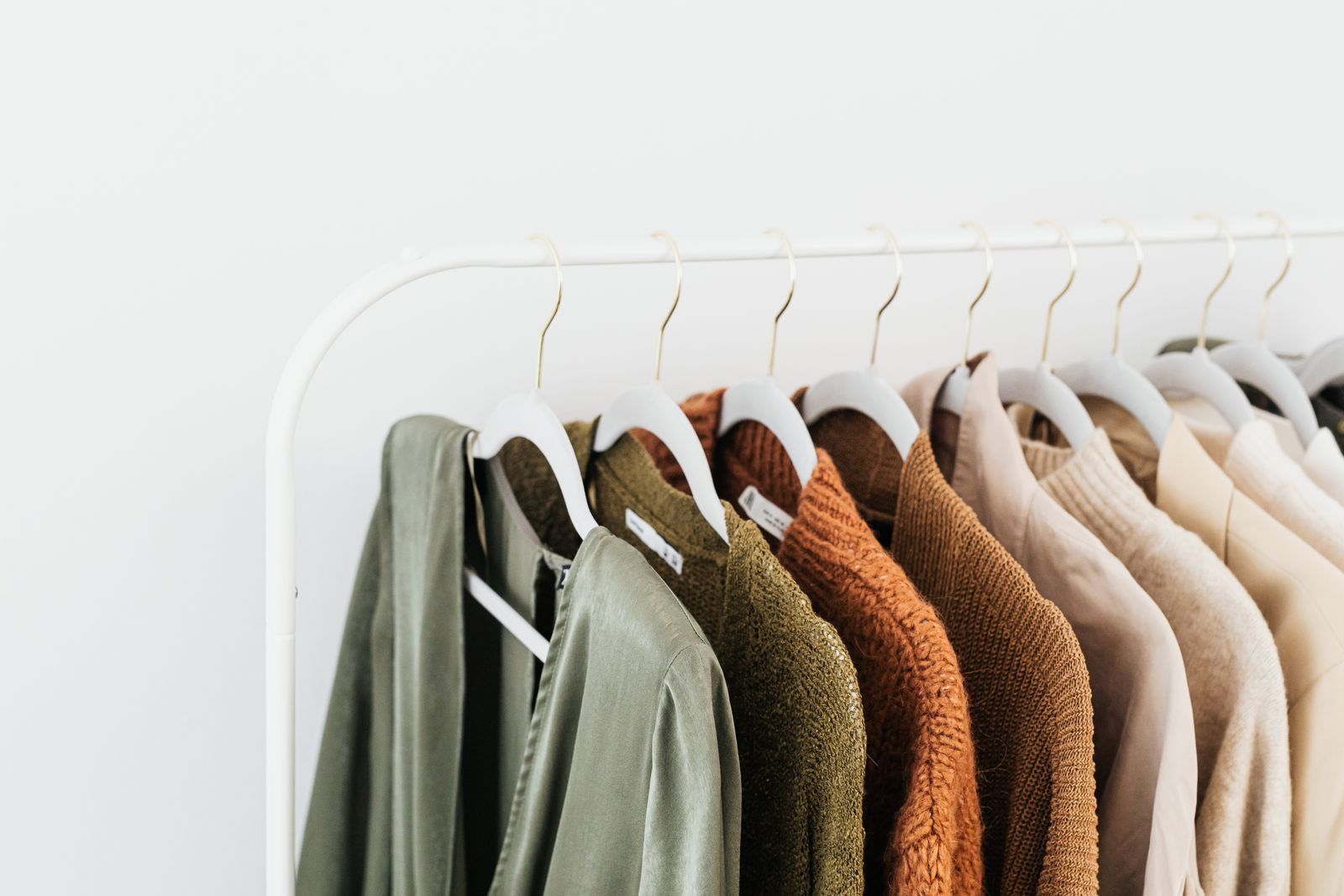 White clothing rack holding a small capsule wardrobe of sage, olive, rust and cream sweaters and shirts on white hangers