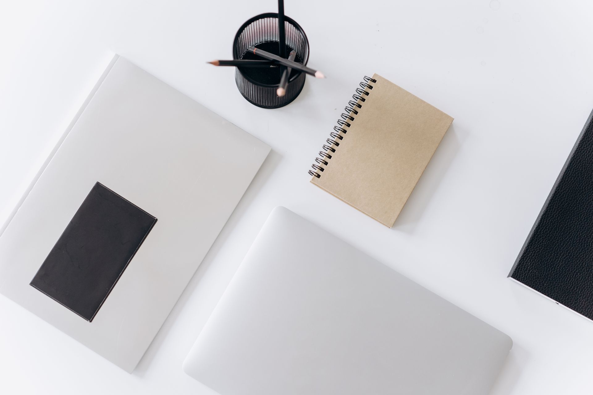 Overhead view of a tidy white desk with a closed laptop, a kraft spiral notebook, a black folder and a mesh cup of pencils