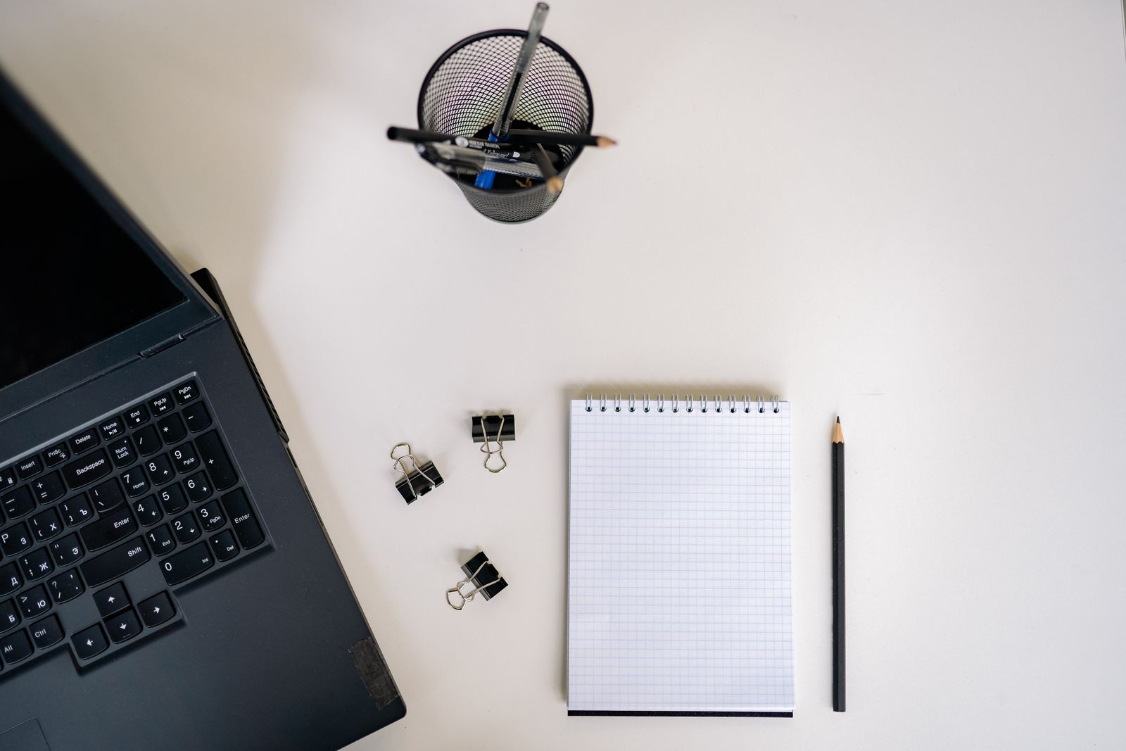 Overhead view of a white desk with an open laptop, a blank grid notepad, a pencil, binder clips and a mesh pen holder