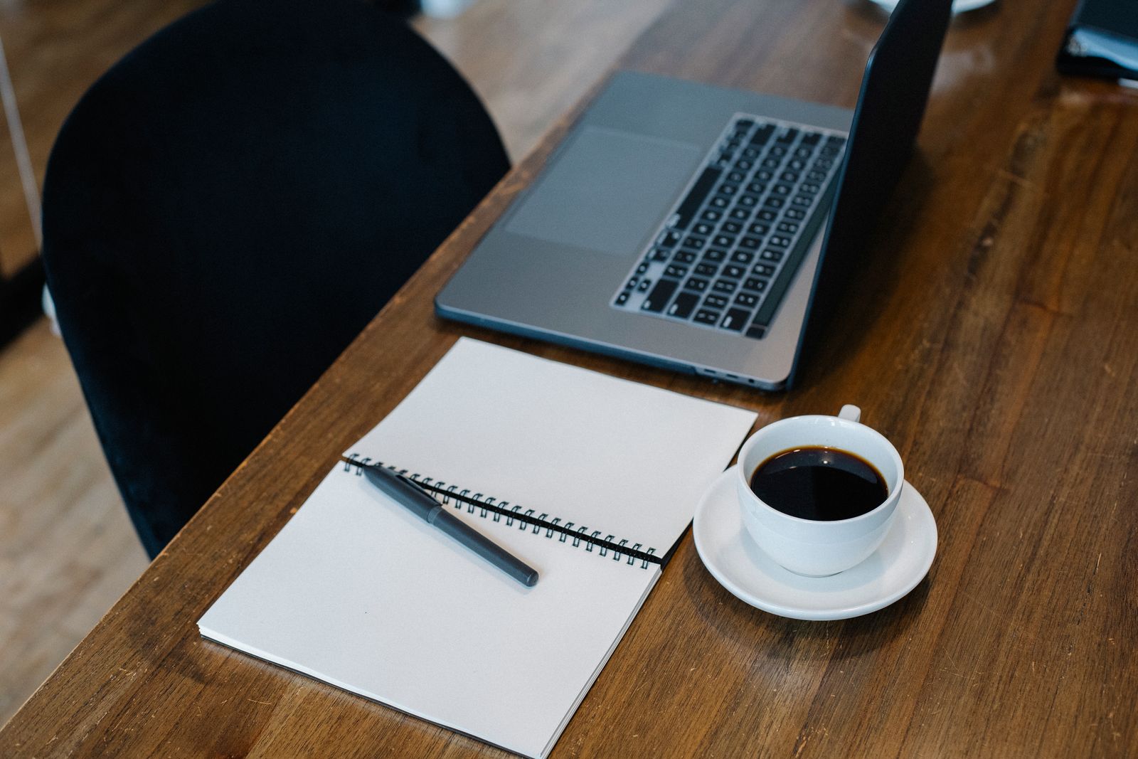 Open laptop on a wooden desk beside a blank spiral notebook, a pen and a cup of black coffee