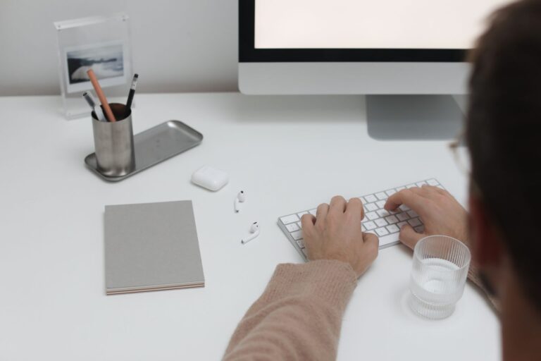 Man typing on a white keyboard at a minimalist white desk with a monitor, pen holder, notebook and glass of water
