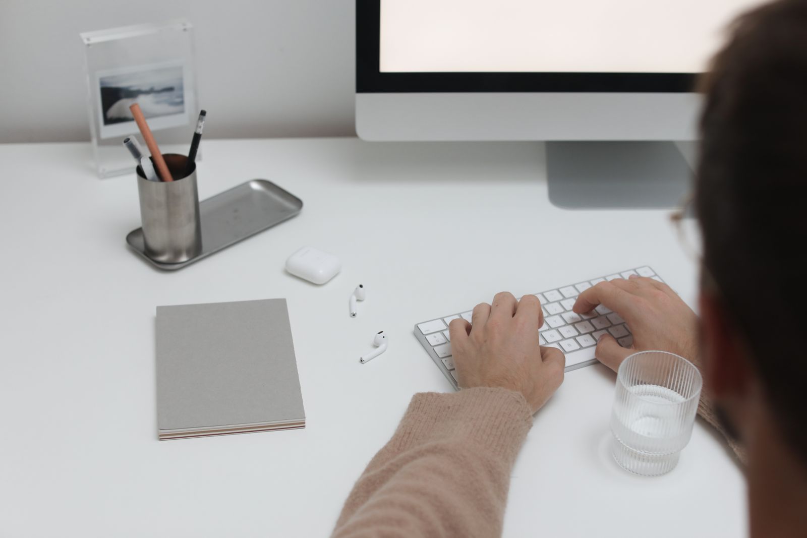 Man typing on a white keyboard at a minimalist white desk with a monitor, pen holder, notebook and glass of water