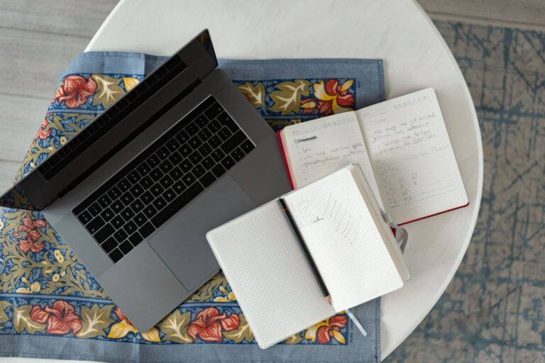 Overhead view of an open laptop beside two open notebooks with handwritten notes and a pencil on a round table