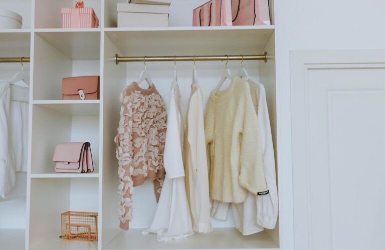 Organized white closet with neutral sweaters and shirts on wooden hangers, shelves holding handbags and storage boxes