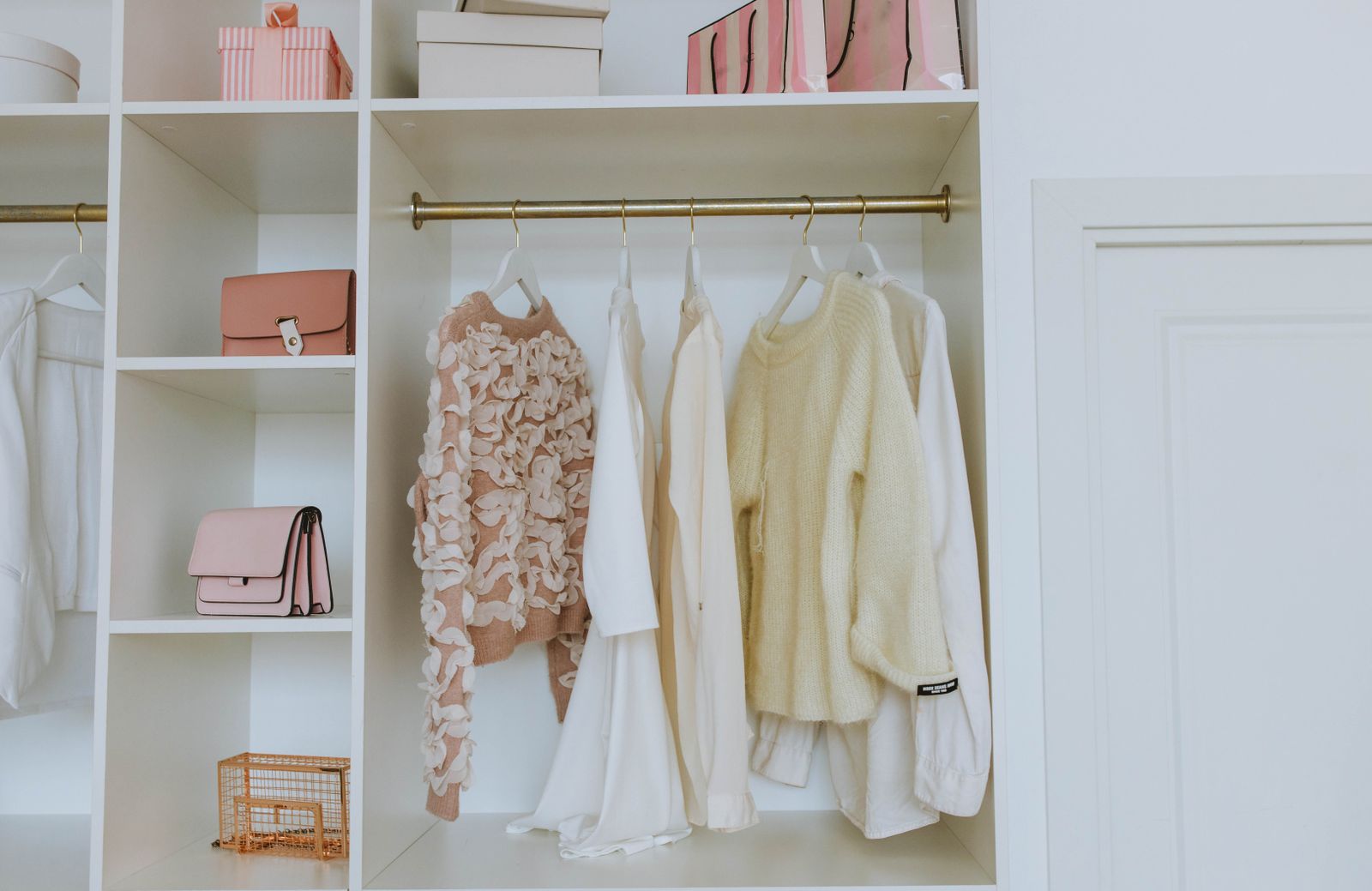 Organized white closet with neutral sweaters and shirts on wooden hangers, shelves holding handbags and storage boxes