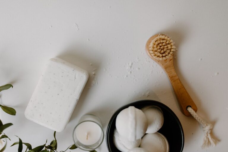 Flat lay of bath essentials on a white surface: a bar of soap, a wooden dry brush, a small candle, bath bombs in a black bowl and green leaves