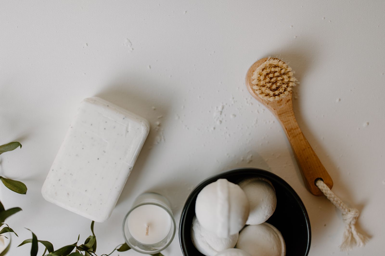 Flat lay of bath essentials on a white surface: a bar of soap, a wooden dry brush, a small candle, bath bombs in a black bowl and green leaves