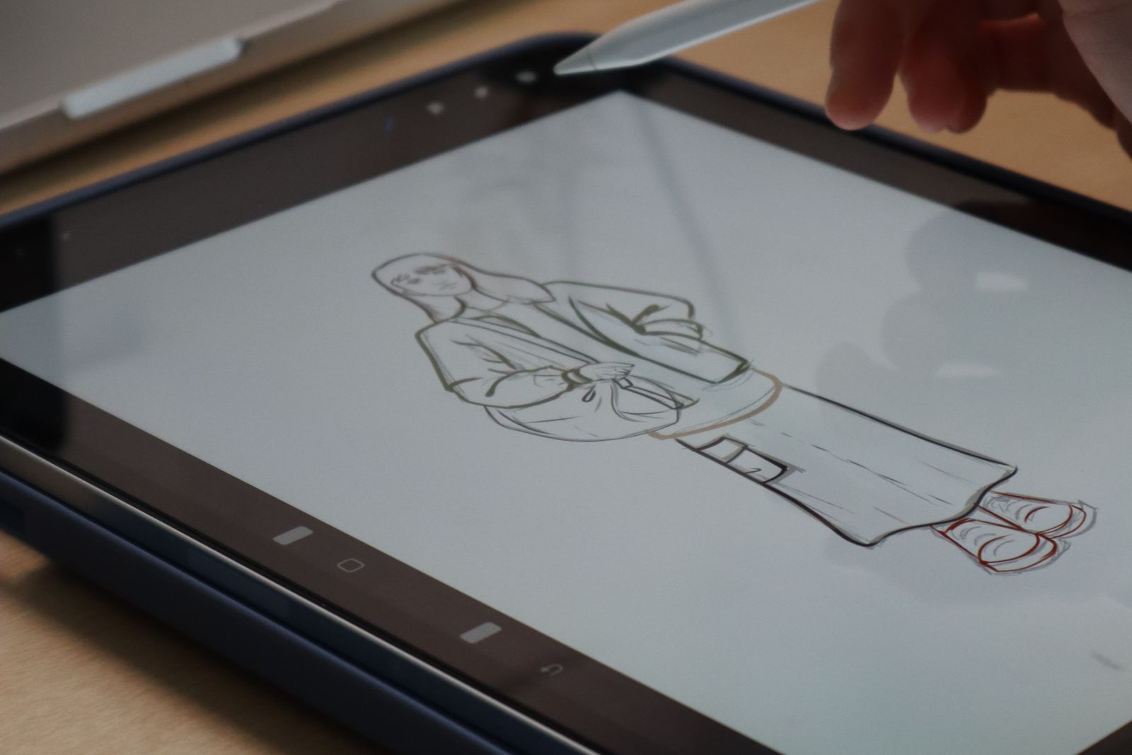 Hand sketching a fashion figure on a tablet with a stylus at a wooden desk beside a laptop